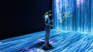 A young person interacts with their smartphone, standing on a glowing floor that projects a dynamic light display, within a dark room illuminated by a large, vivid digital art installation.